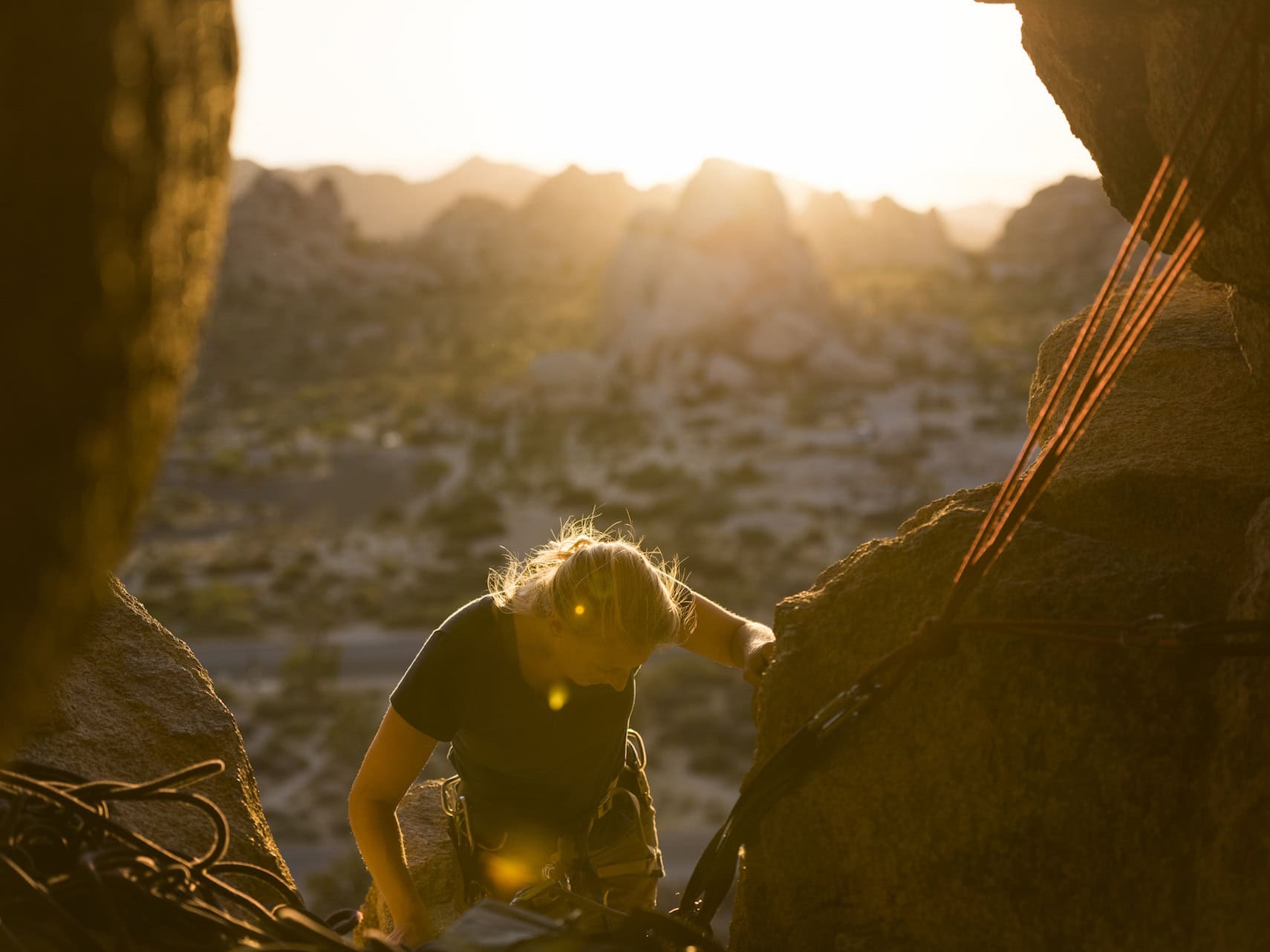 A woman climbs between angled slants of rock while the sun rises behind her, illuminating the tree-dotted ground far below.