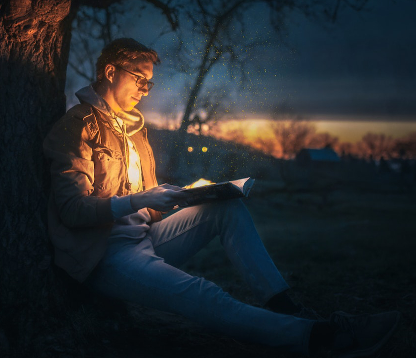 A man sitting under a tree reading, while light spills up like golden sparks from the pages, illuminating his face.