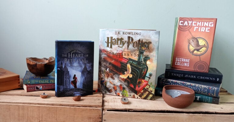 An array of fantasy novels are set up on pale wooden crates in front of a pale green wall, arfully arranged among clay candles, an olive wood bowl, a maple box, and miniscule walnut candles