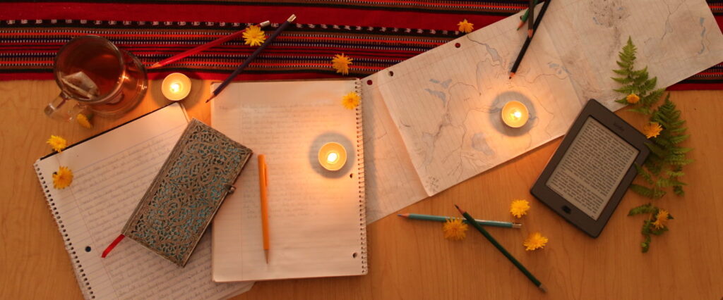 A collection of notebooks, hand-drawn maps, tea candles, a kindle, and colored pencils spread across a table adorned with a red table runner and dandelion flowers.