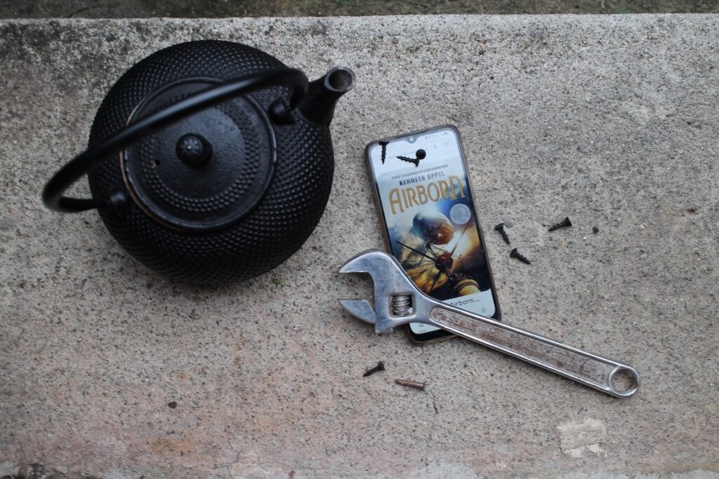 A cast iron teapot, an adjustable wrench, and black screws are arranged on concrete around a phone with the cover of book on the screen. It shows an airship and the title "Airborne" by Kenneth Oppel, a steampunk fantasy for middle grade readers.