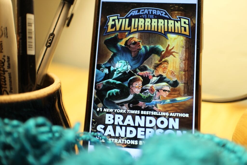 Several exagerrated figures wearing sunglasses and enormous swords race across the screen of a phone, showing the cover of Alcatraz versus the Evil Librarians by Brandon Sanderson, a hilarious comic fantasy for middle-grade readers (pre-teen).