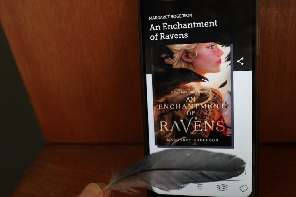 A phone screen shows a book cover of a crow sitting on a girl's shoulder.