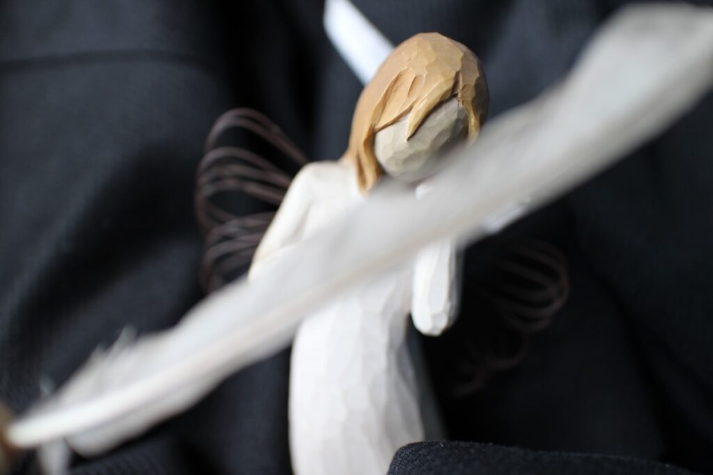 A carved wooden angel on a black background, with a feather cutting diagonally across the foreground. It is meant to portray Hopepunks rejection of cynicism.
