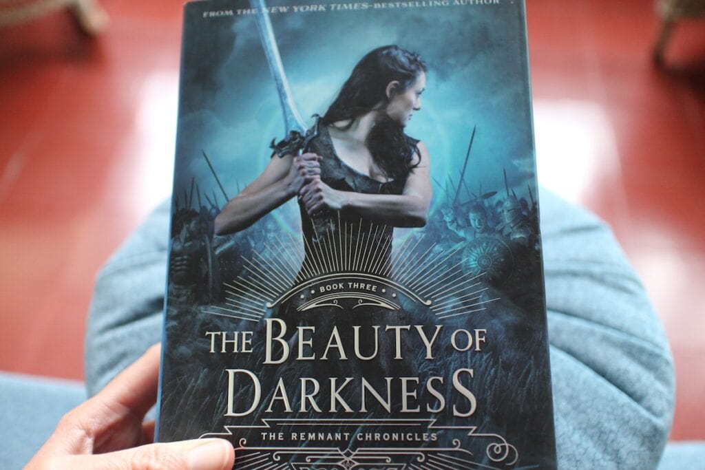A woman in a black dress lifts a sword dramatically. This is the cover of "The Beauty of Darkness" the final book in "The Remnant Chronicles" by Mary E. Pearson. This is as example of medieval fantasy.