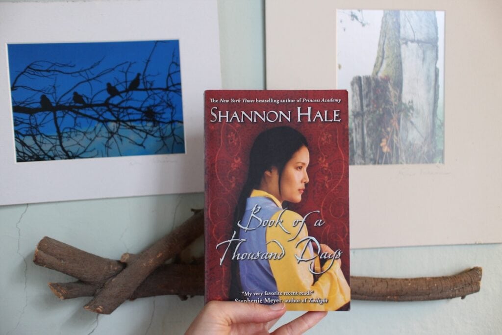 A hand holds a book in front of a branch of wood. The cover shows a black-haired girl with Mongolian features clutches a notebook. This is "Book of a Thousand Days" by Shannon Hale, a fairy-tale fantasy.