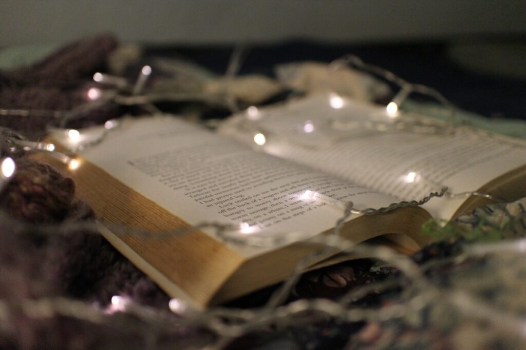 A book open on blankets and surrounded by fairy lights. This is an example of cozy fantasy.
