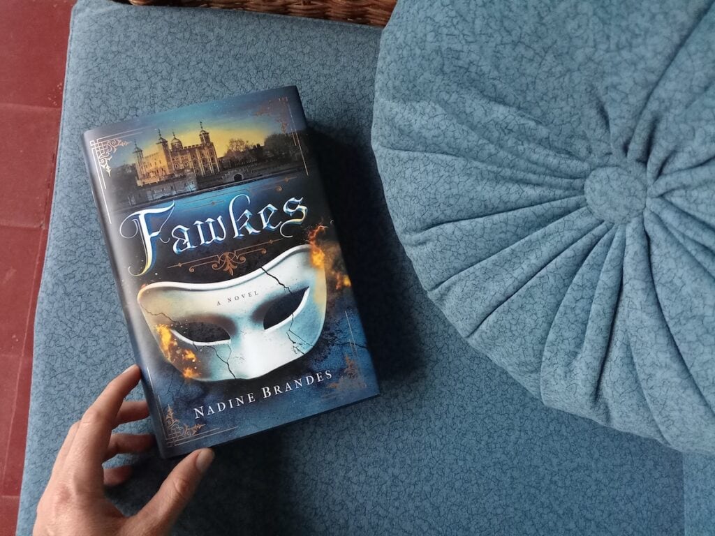 A blue book with a white crackled mask and glints of fire sits on a blue couch. The title reads "Fawkes" by Nadine Brandes, an excellent example of a historical fantasy.