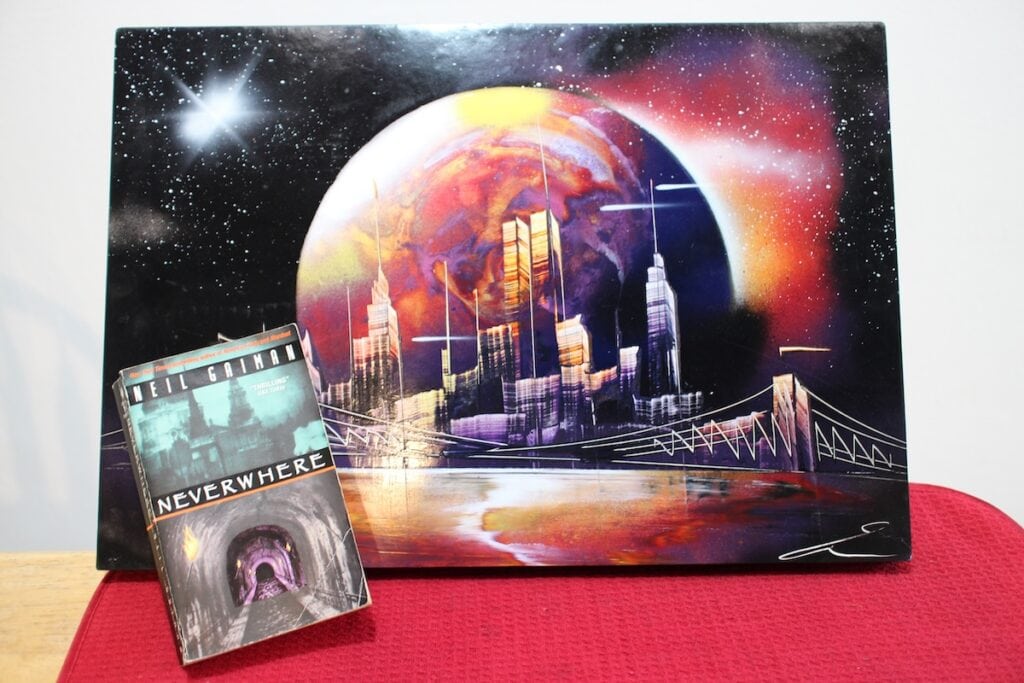 A book showing a picture of London and a train track disappearing underground sits in front of a dramatic painting of yellow and purple planet with a city of skyscrapers rising in front of it. Neverwhere is a great example of Urban Fantasy.