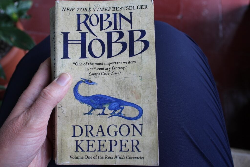 A woman holds a book with a blue dragon on the cover on her lap. The title reads "Dragon Keeper - The Rainwilds Chronicles."