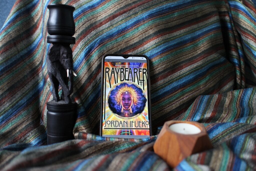 A carved blackwood elephant candlestick, a phone, and a tea candle inside a wooden holder are arranged in front of a blue, green, and gold scarf from Tanzania. The phone screen shows the cover image of "Raybearer" by Jordan Ifueko—an epic Afro-fantasy novel.