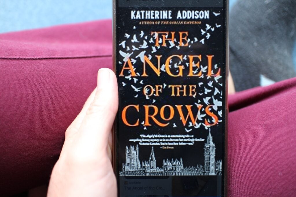 A woman holds a phone with a black book cover dotted with white crows above a stylized city of London. The text reads "The Angel of the Crows," a gaslamp fantasy by Katherine Addison.