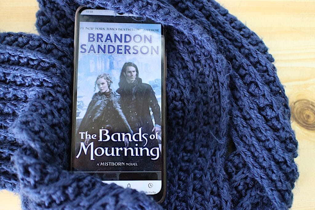 A man and woman stand outdoors in a snowy environment. It is the book cover to Bands of Mourning by Brandon Sanderson.