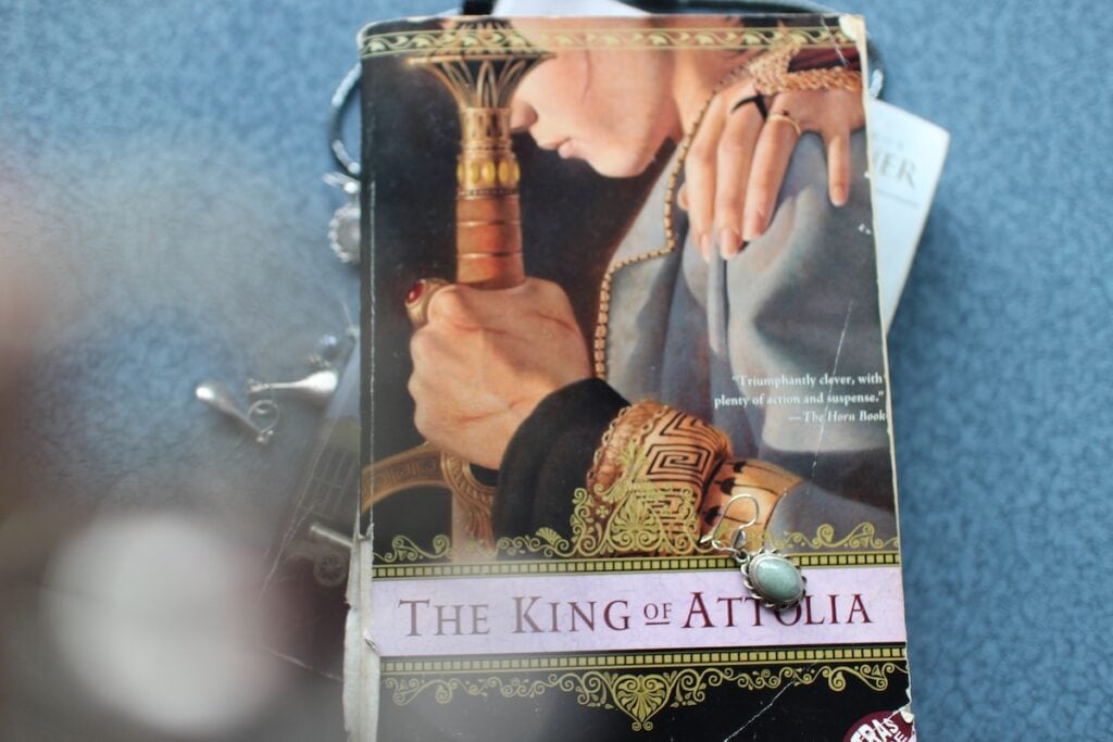 Jewelry drapes across the cover of a book showing the profile of young man with a scarred face gripping a sword. This is one of my favorite political fantasy books.