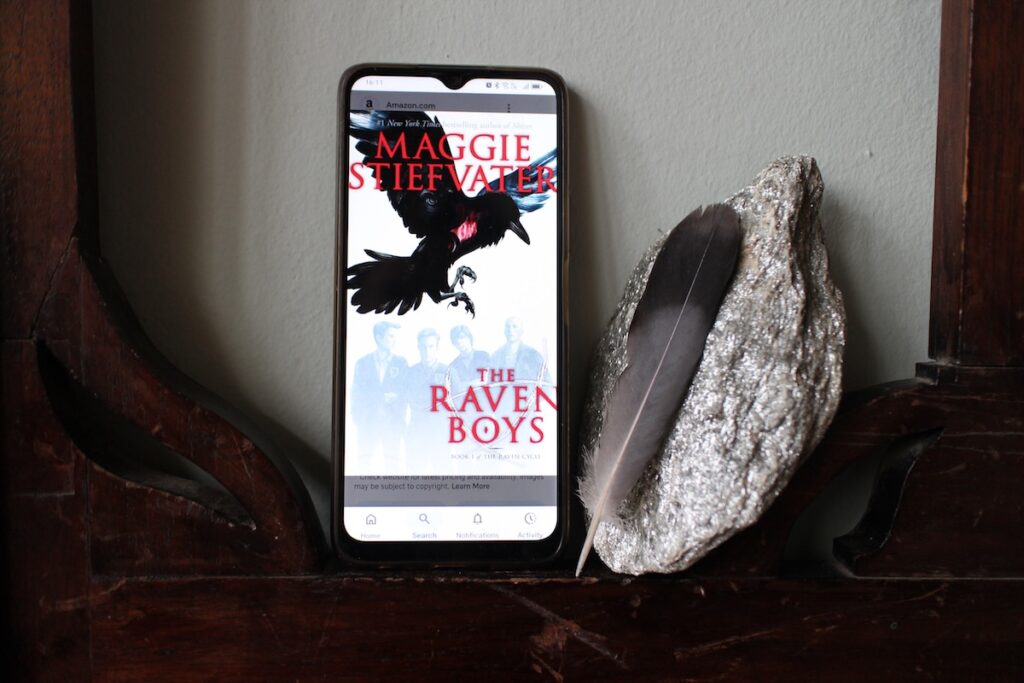 A phone sits between curved slats of wood between a glittering mica-encrusted stone and a gray and black feather. On the screen is the cover image of "The Raven Boys" by Maggie Stiefvater. It shows a crow, legs extended toward four grayed-out teen boys wearing matching jackets. The Raven Boys is a great example of Low Fantasy
