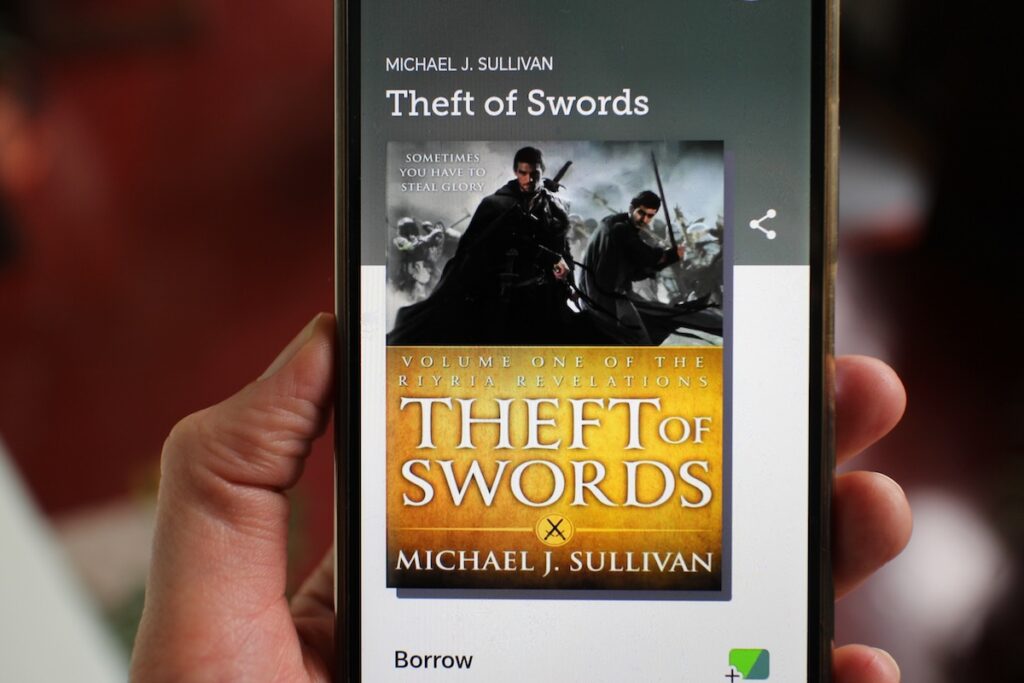 Two cloaked figures with swords appear on the screen of a phone above the title "Theft of Swords." This is a fantastic heist novel in the roguish fantasy subgenre.