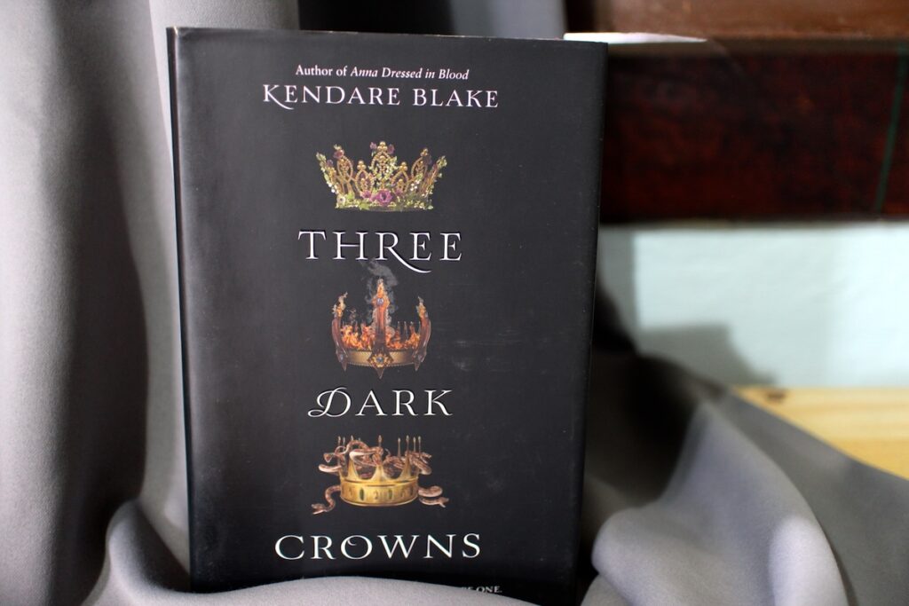 A black book with three different crowns arrayed down the center, one with plants, one with snakes, and one with fire. "Three Dark Crowns" by Kendare Blake is a great example of the deadly competition trope.