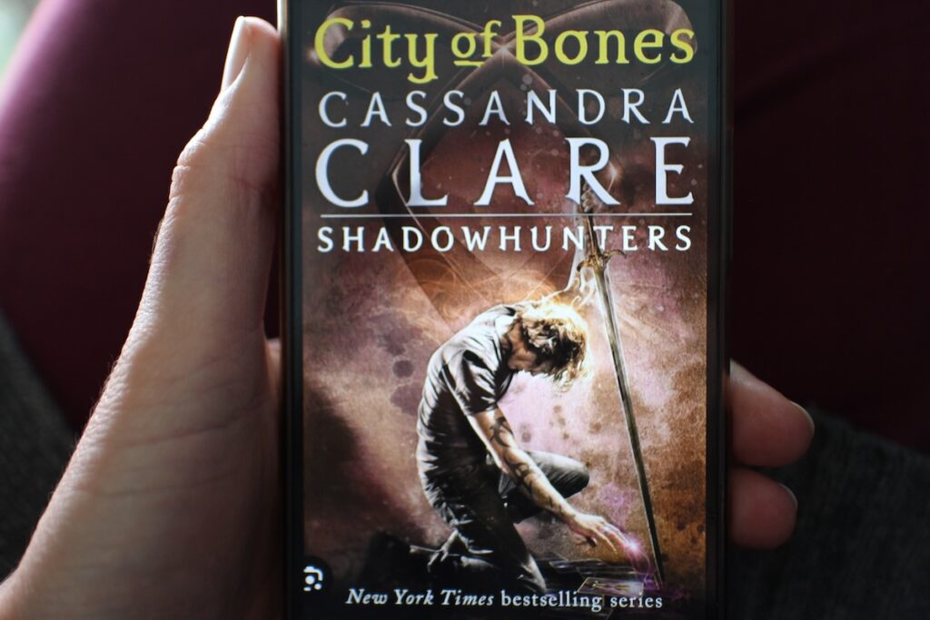 A woman holds a cell phone with a book cover on the screen. It shows a young man with runes pained on his arms surrounded by mythical lights. "City of Bones" by Cassandra Clare is a paranormal fantasy set in New York City.