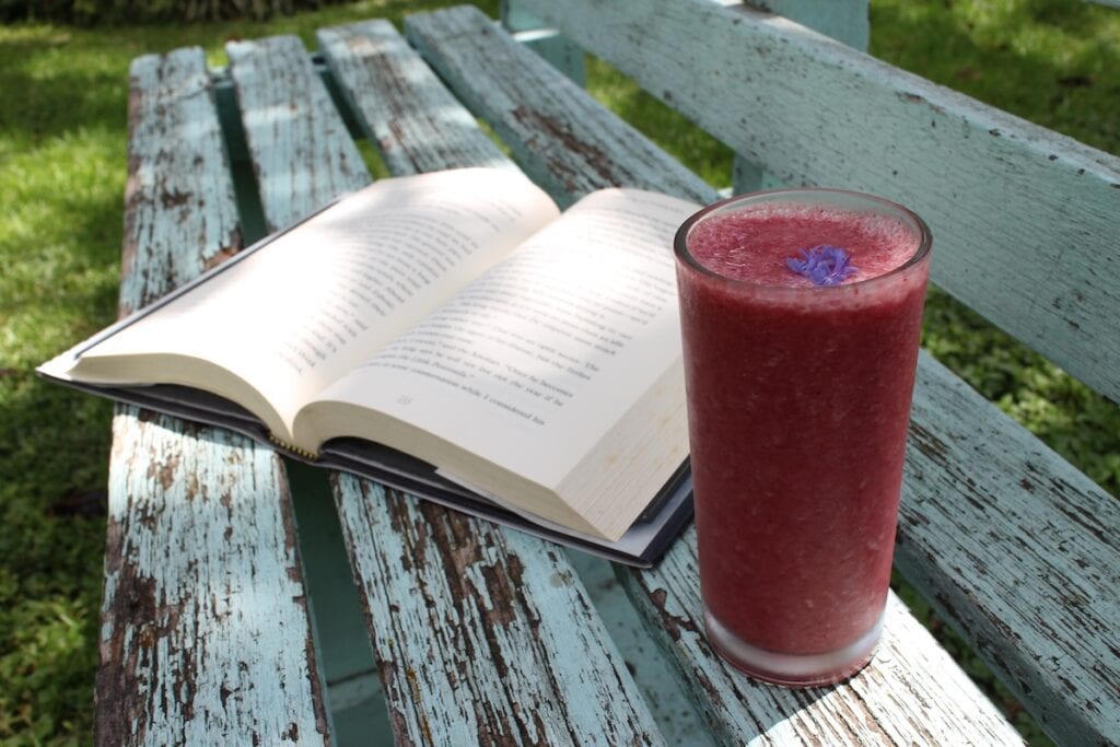 A novel lies open next to a berry smoothie on an old wooden bench with natural wood showing through aging blue paint